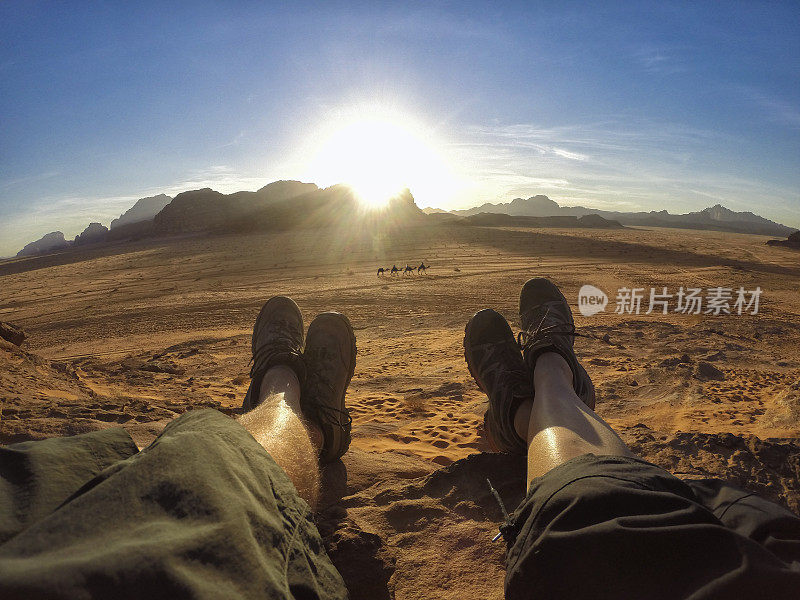 一对夫妇的脚休息在红色的砂岩上，看着骆驼通过Wadi Rum沙漠，约旦